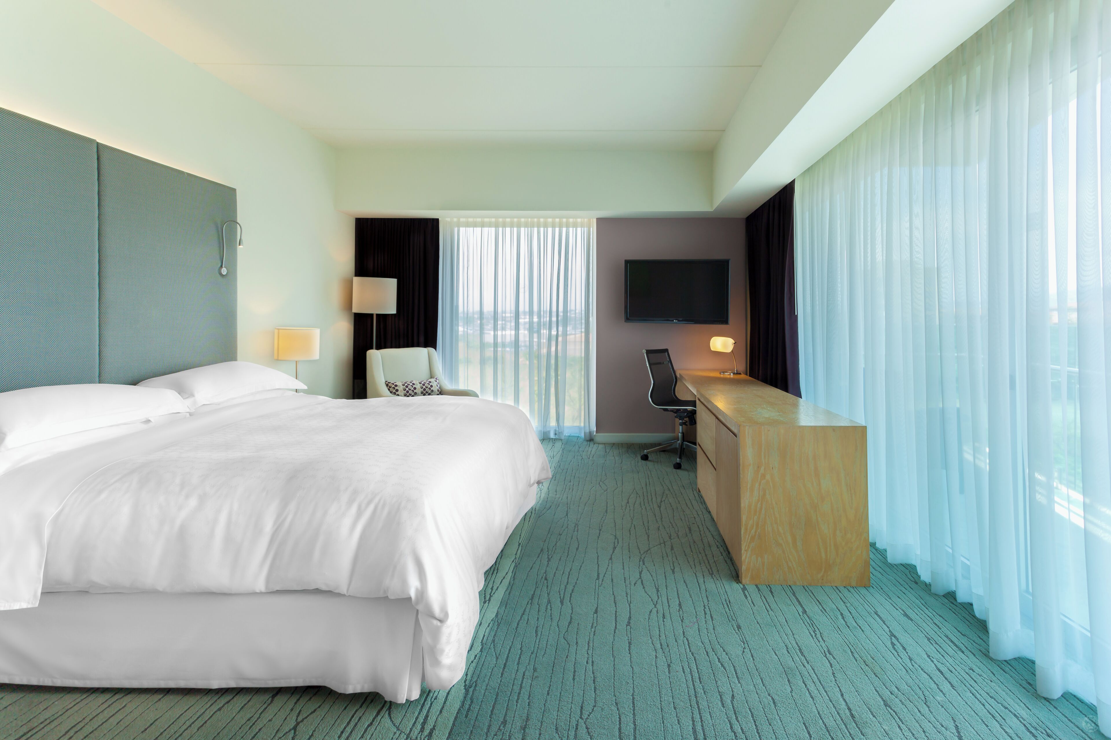A hotel room with a large bed, a desk, a chair, a TV, and a view of the city through the window.