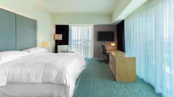 A hotel room with a large bed, a desk, a chair, a TV, and a view of the city through the window.