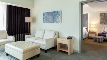 A hotel room with a sofa, ottoman, a small wooden side table with a phone, a painting on the wall, and a view into the adjacent room.