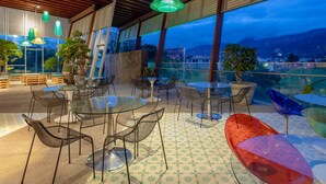 An outdoor patio with round tables and chairs, large windows with a city view, and hanging lamps.