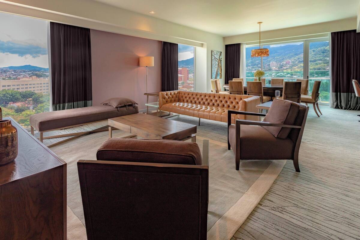 A modern hotel room with a large sofa, armchairs, a dining table, and a view of a cityscape.