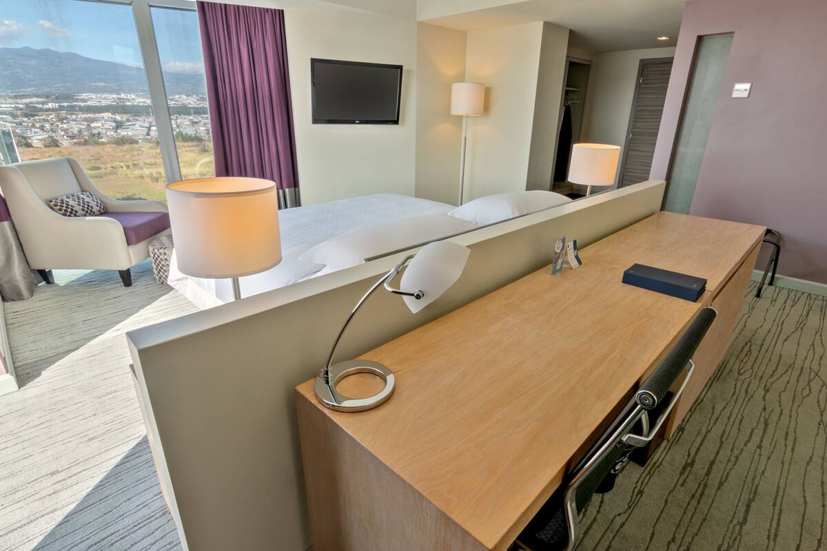 A hotel room with a large bed, a desk, a chair, a lamp, and a TV.