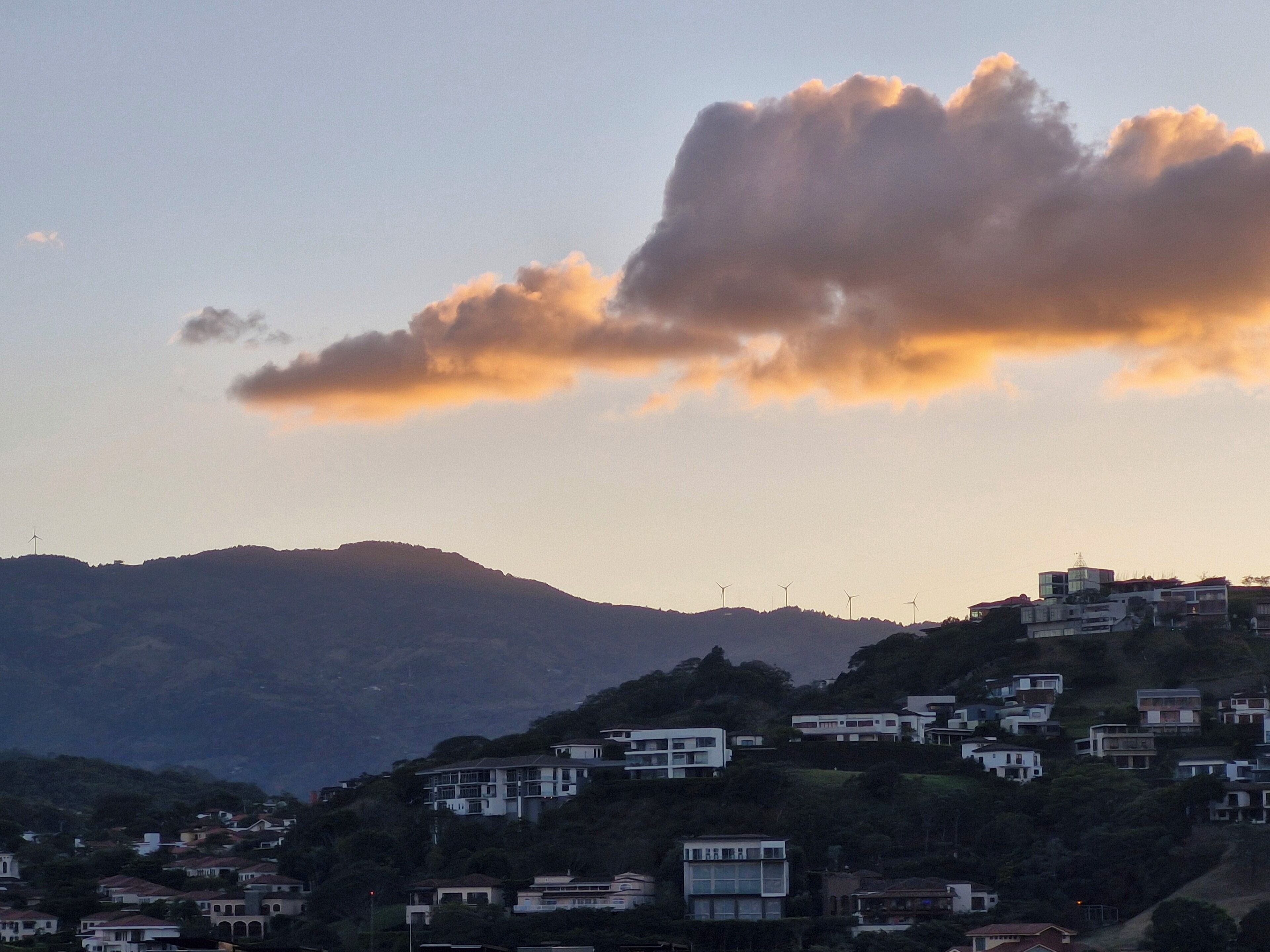 Un quartier résidentiel avec des maisons construites sur une colline, un nuage imposant dans le ciel et des éoliennes visibles au loin.