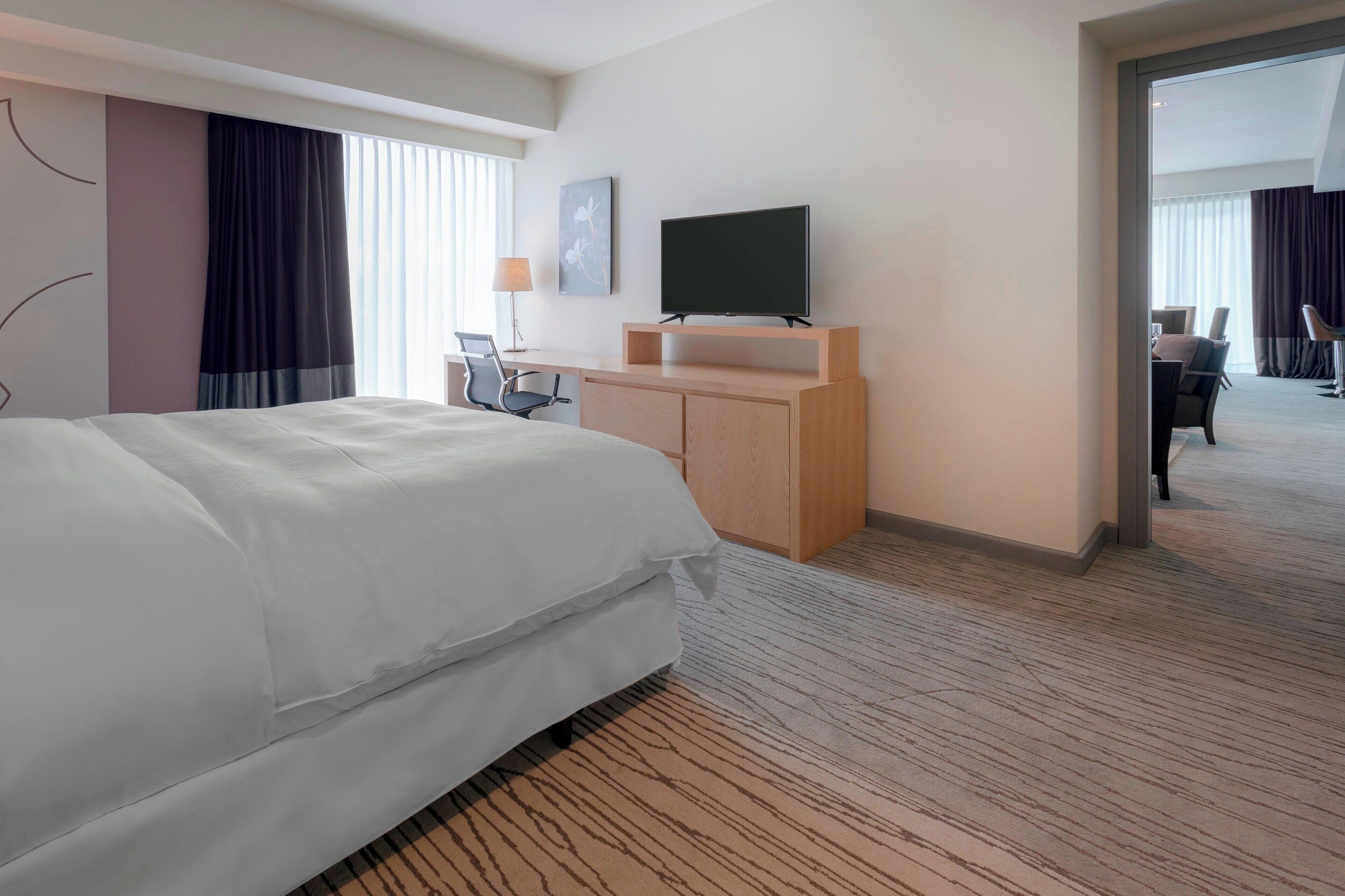 A hotel room with a large bed, a desk with a chair, a flat-screen TV, and a view of the hallway.