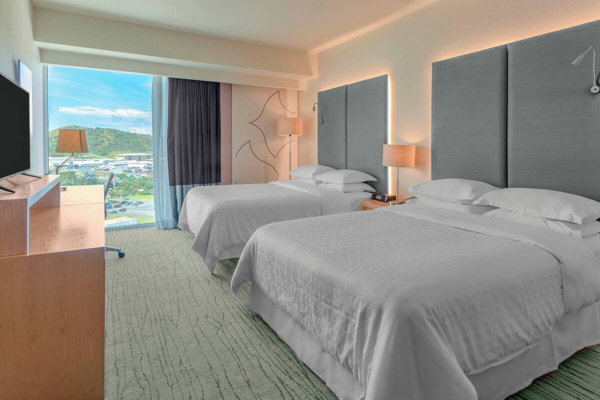 A hotel room with two beds, a desk, and a large window offering a city view.