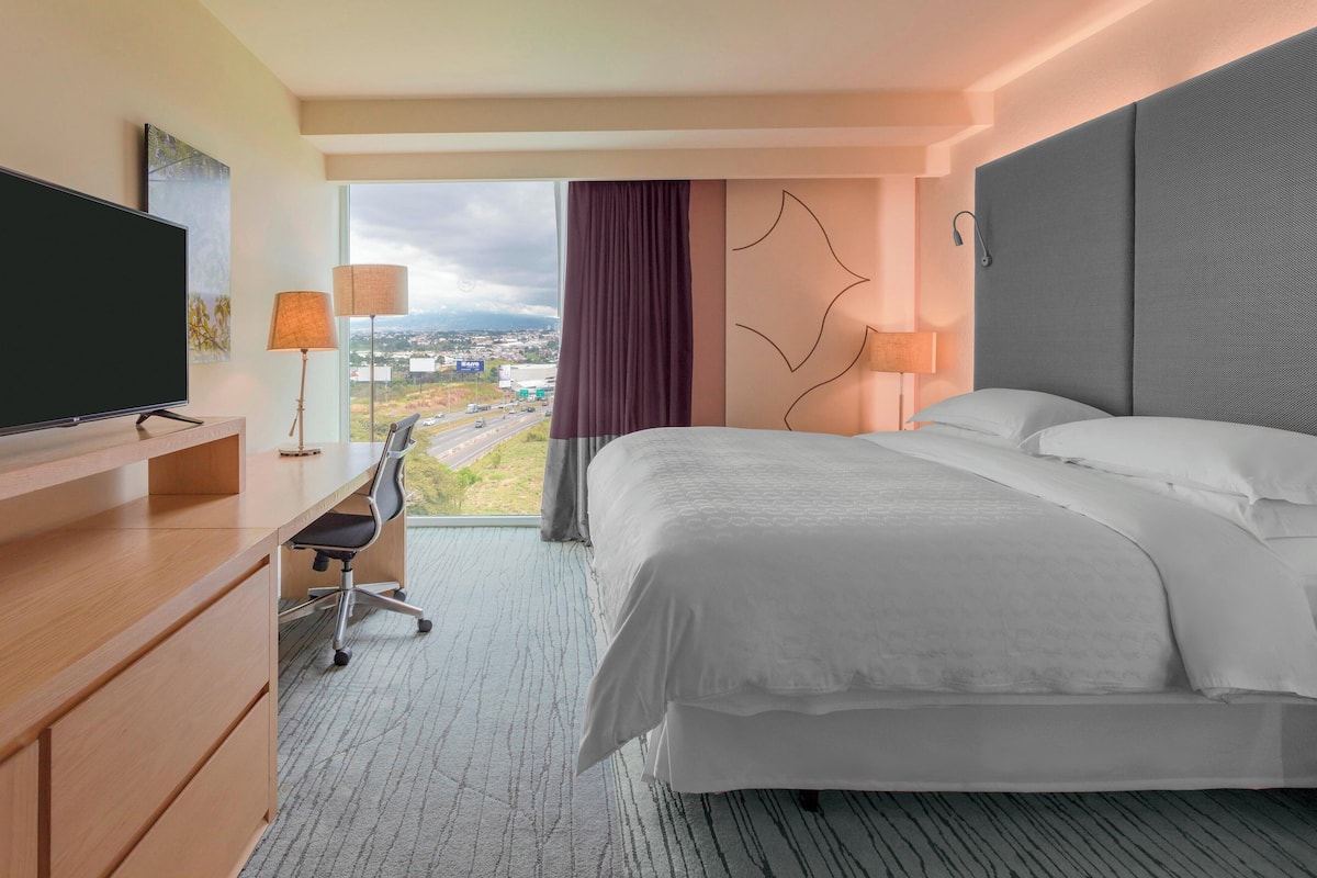 A hotel room with a large bed, a desk with a chair, a flat-screen TV, and a view of a cityscape through a large window.