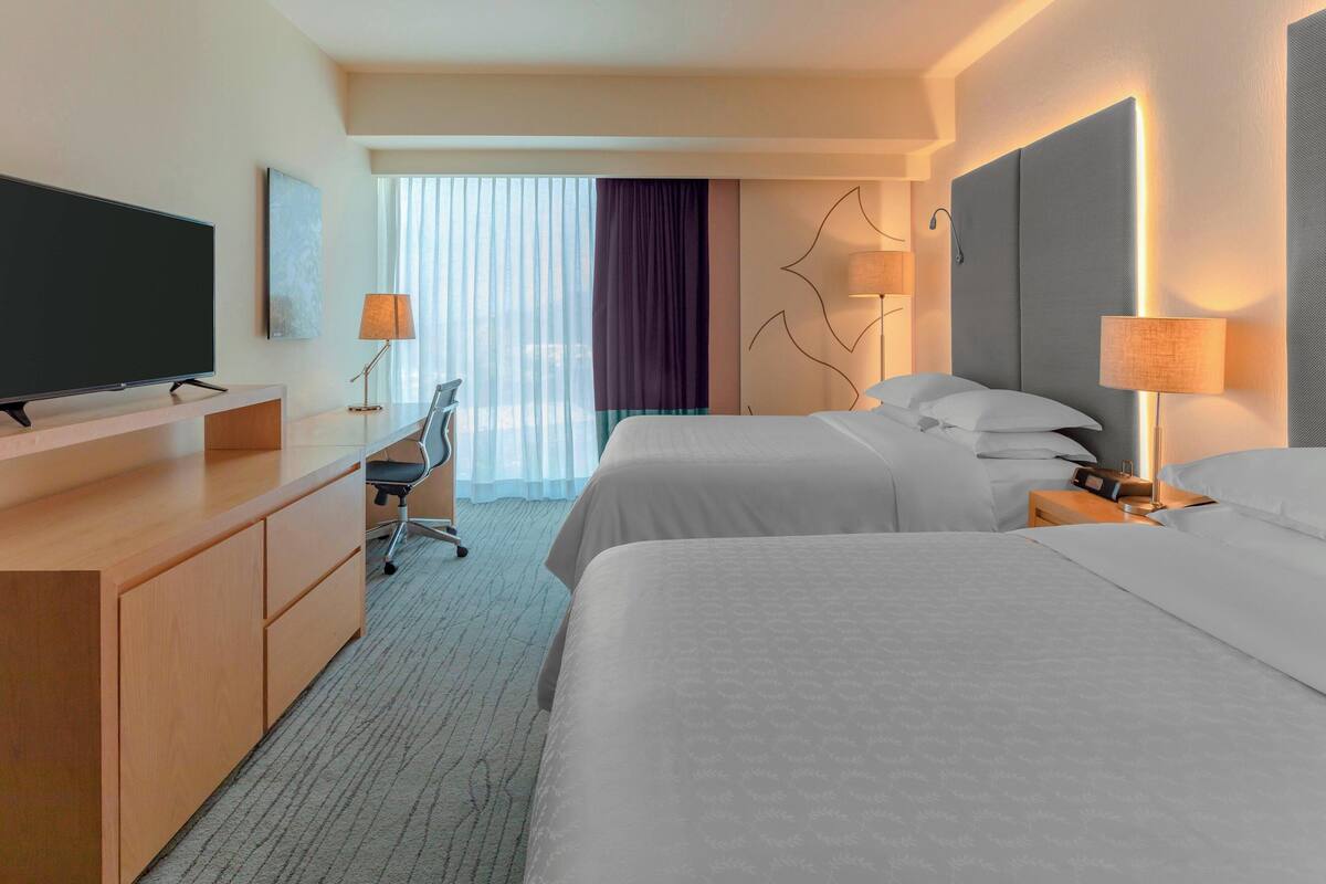 A hotel room with two beds, a desk with a flat-screen TV, a chair, and a large window with curtains.