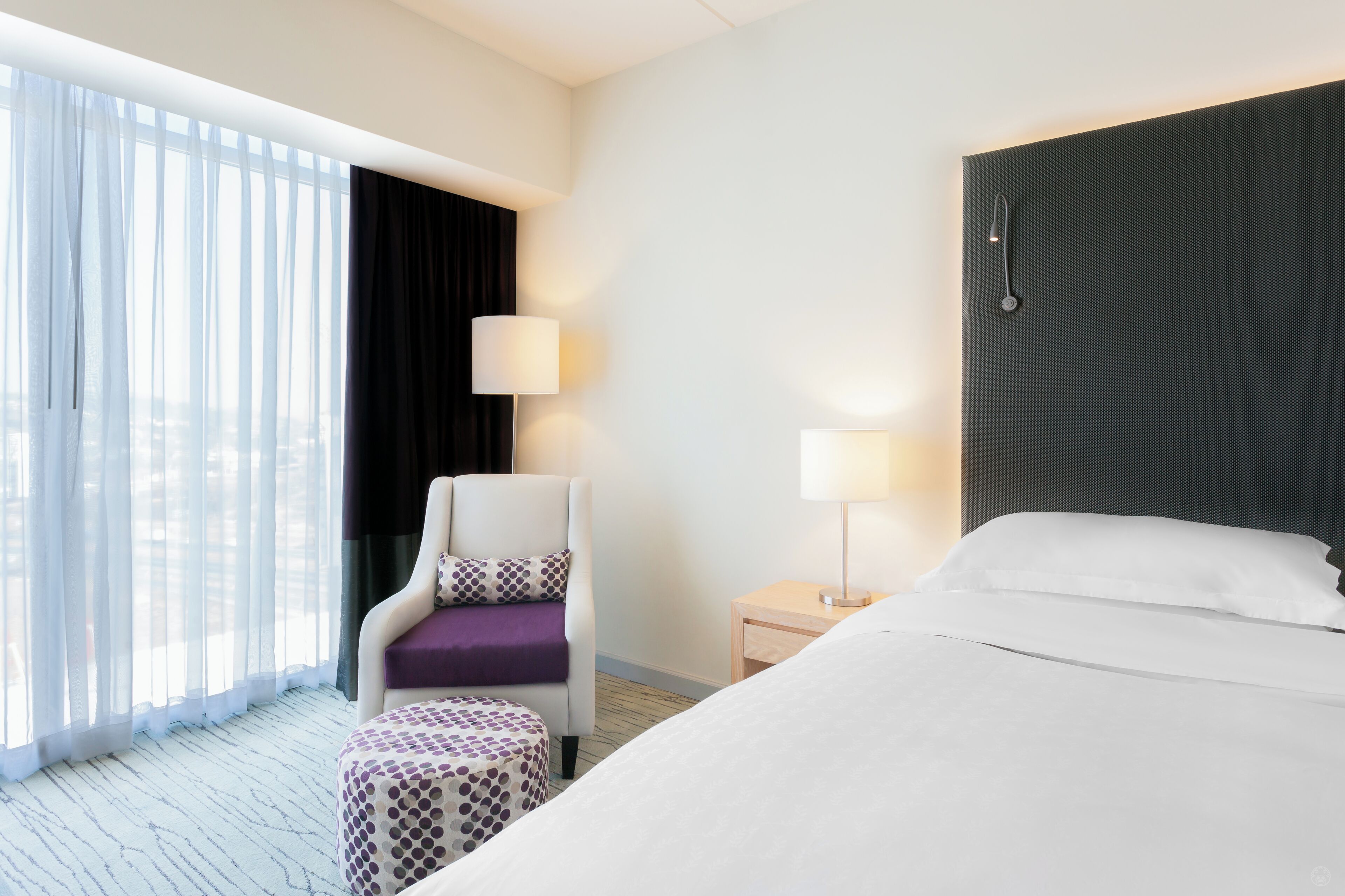 A hotel room with a large bed, a chair with a purple cushion, a small table with a lamp, and a view of the outdoors through sheer curtains.
