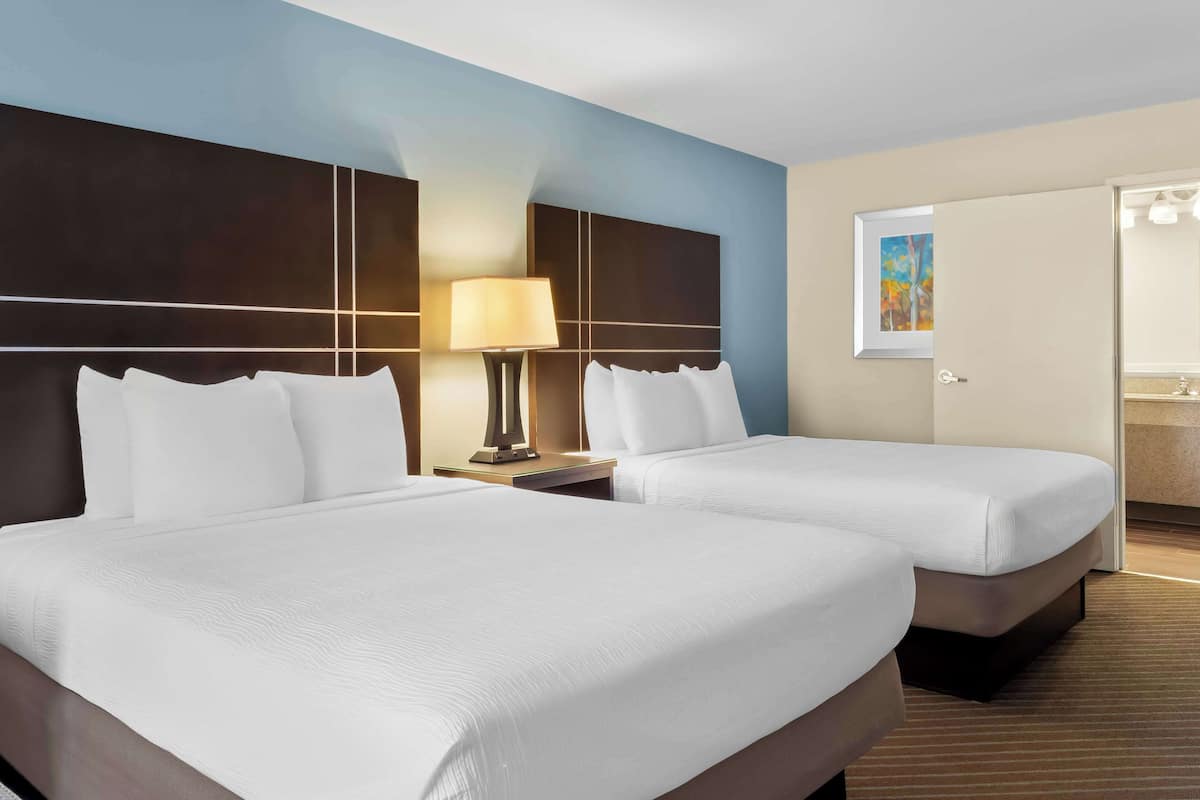A hotel room with two beds, a nightstand with a lamp, a painting on the wall, and a bathroom door.
