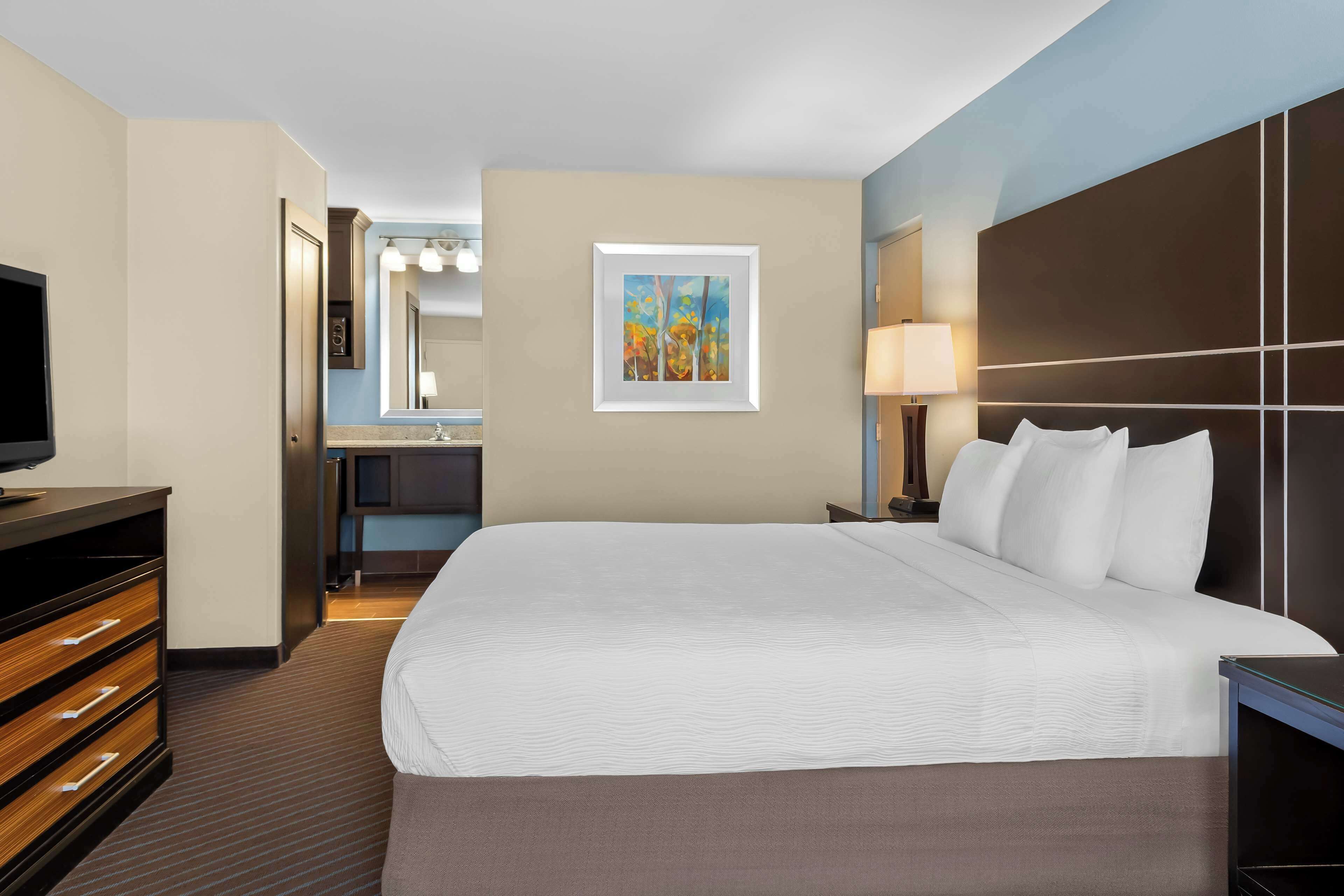 Un cuarto de hotel con una cama grande, un televisor, un escritorio con una lámpara, un baño con un espejo y un cuadro en la pared.