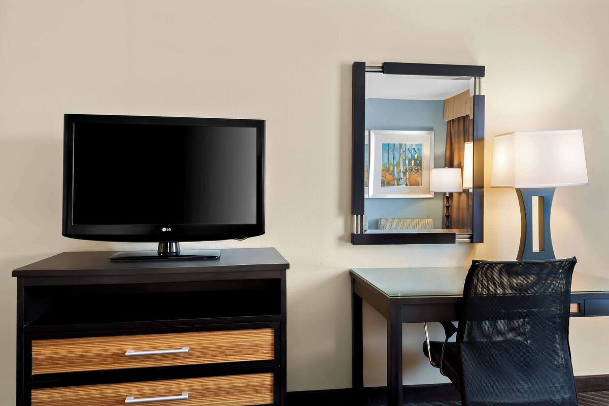 A hotel room with a flat-screen TV on a black TV stand, a desk with a chair, a lamp, and a mirror reflecting the room.