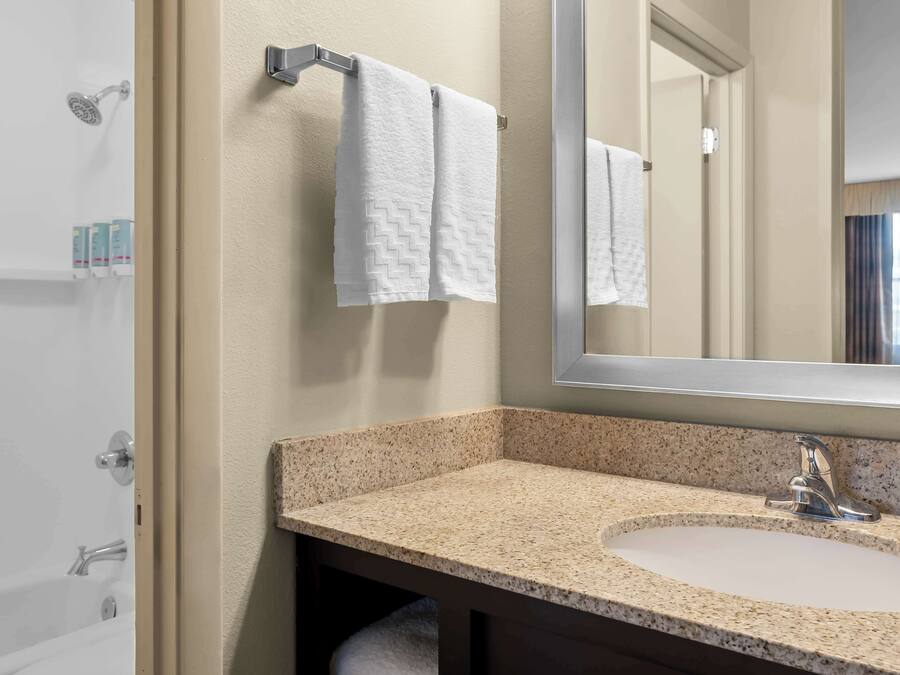Une salle de bain avec un lavabo doté d’un plan de travail en granit, un grand miroir et deux serviettes blanches suspendues à un porte-serviettes.