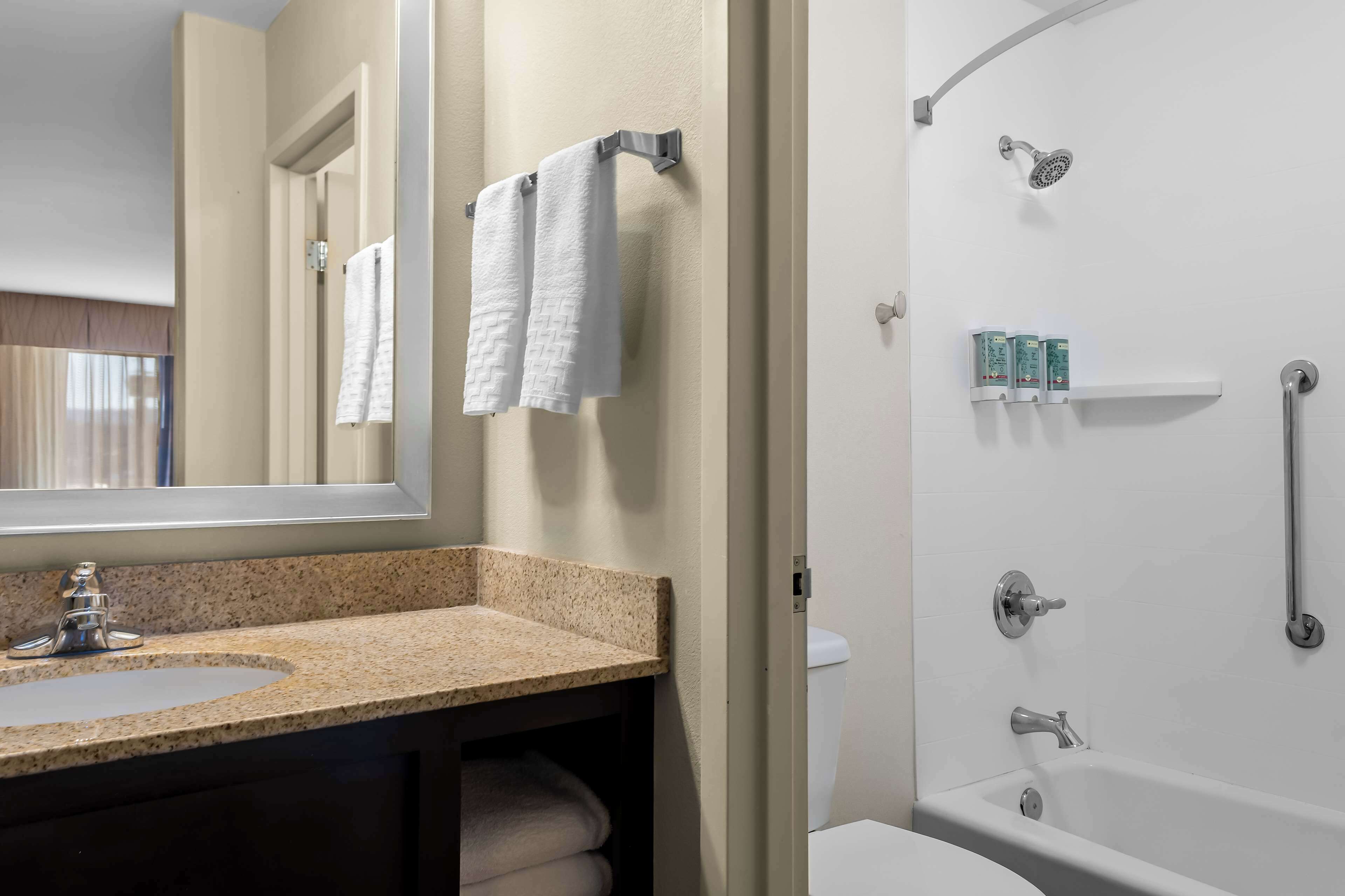 A bathroom with a granite countertop sink, a mirror, two white towels, a shower with a handheld showerhead, and a toilet.