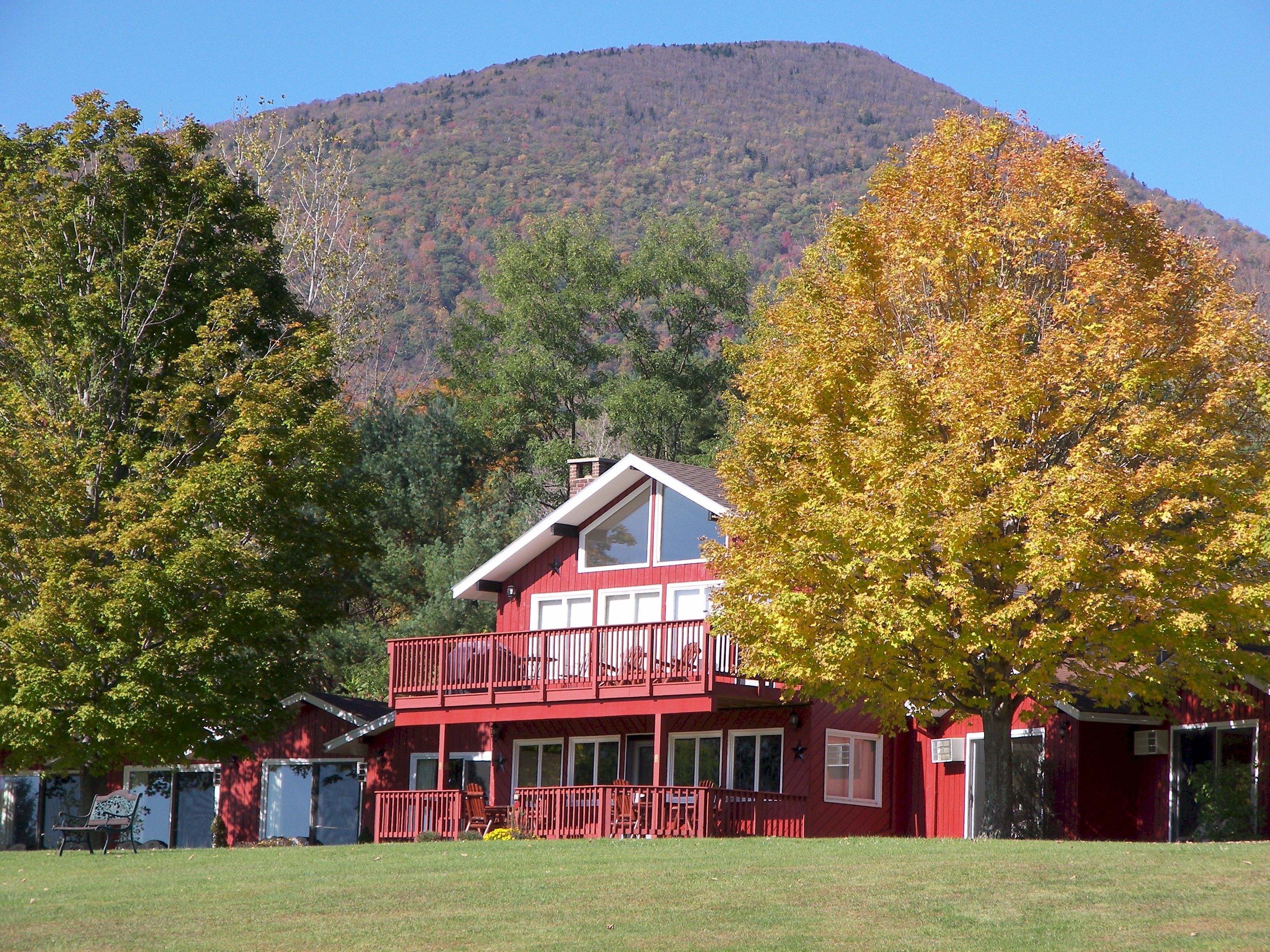 A two-story red building with a balcony, surrounded by trees with autumn foliage, set against a backdrop of a hill.