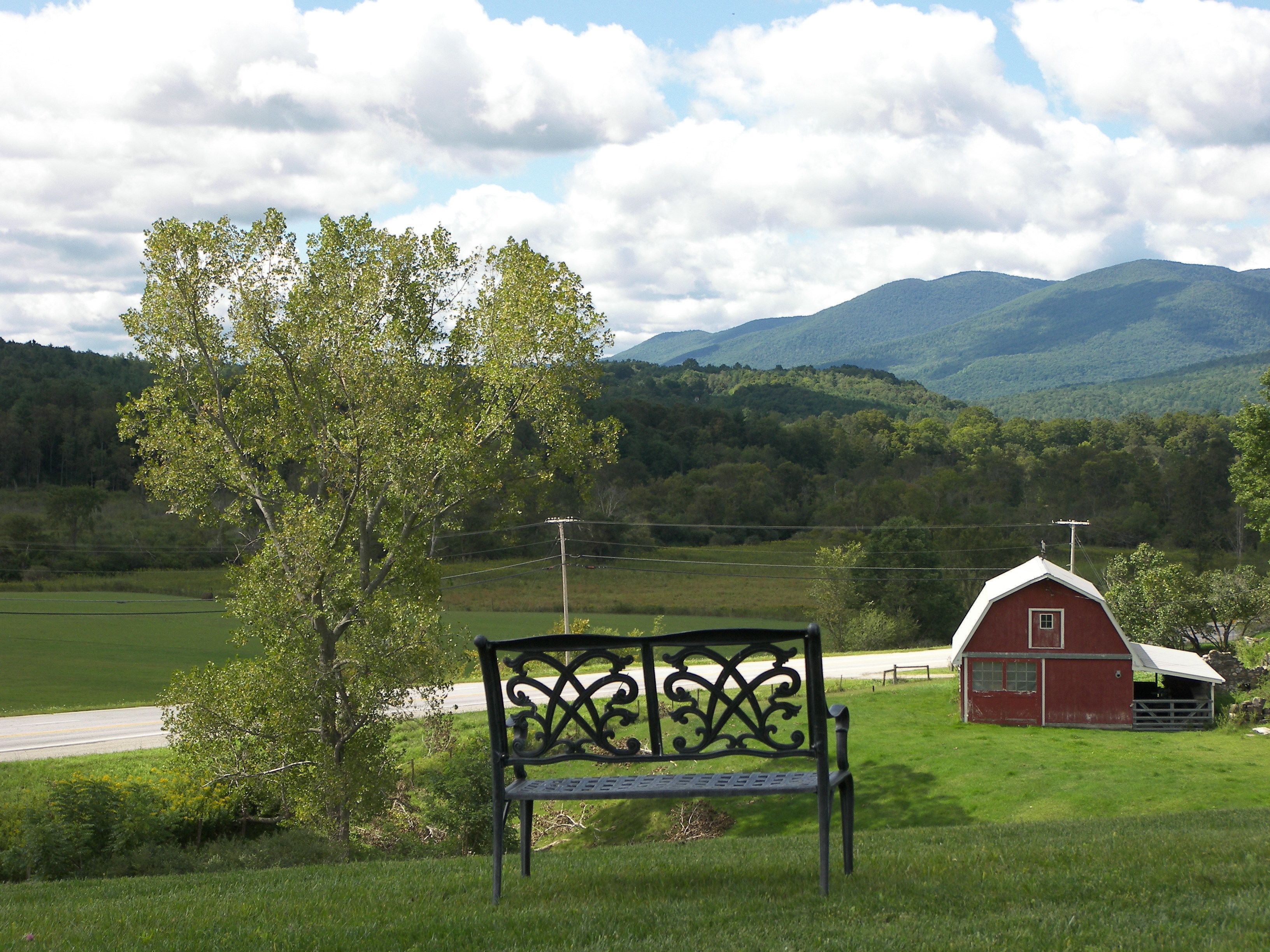 A bench in a grassy field with a red barn and mountains in the background.