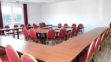 Un salón de conferencias con varias mesas y sillas rojas, ventanas amplias y un proyector de techo.