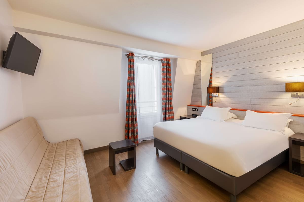 A hotel room with a bed, a sofa, a TV mounted on the wall, a window with curtains, and a bedside table.