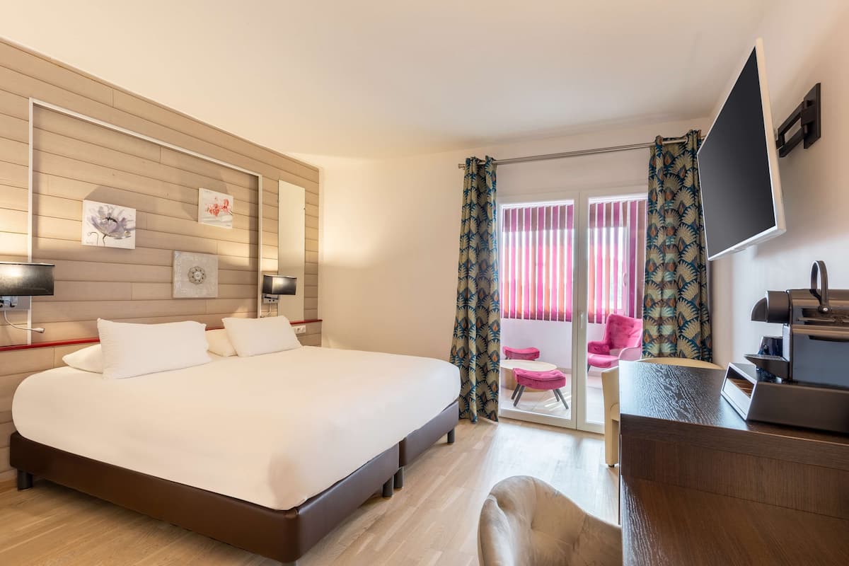 A hotel room with a large bed, a desk with a printer, a TV mounted on the wall, and a sliding glass door leading to a balcony with a pink chair.