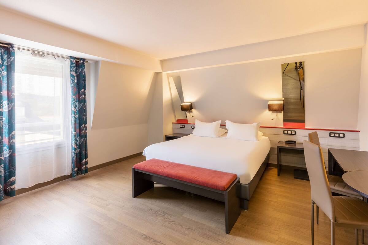 A hotel room with a bed, a desk, a chair, a bench, and a large window with curtains.