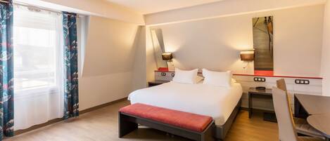 Una habitación de hotel con cama, escritorio, silla, banco y un amplio ventanal con cortinas.
