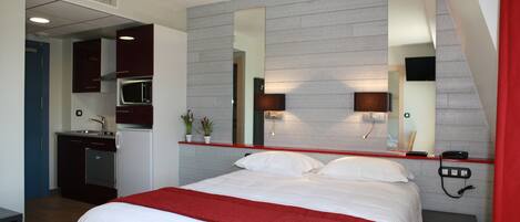 Una habitación de hotel con una cama, un muro de acento rojo, una kitchenette y un espejo grande.