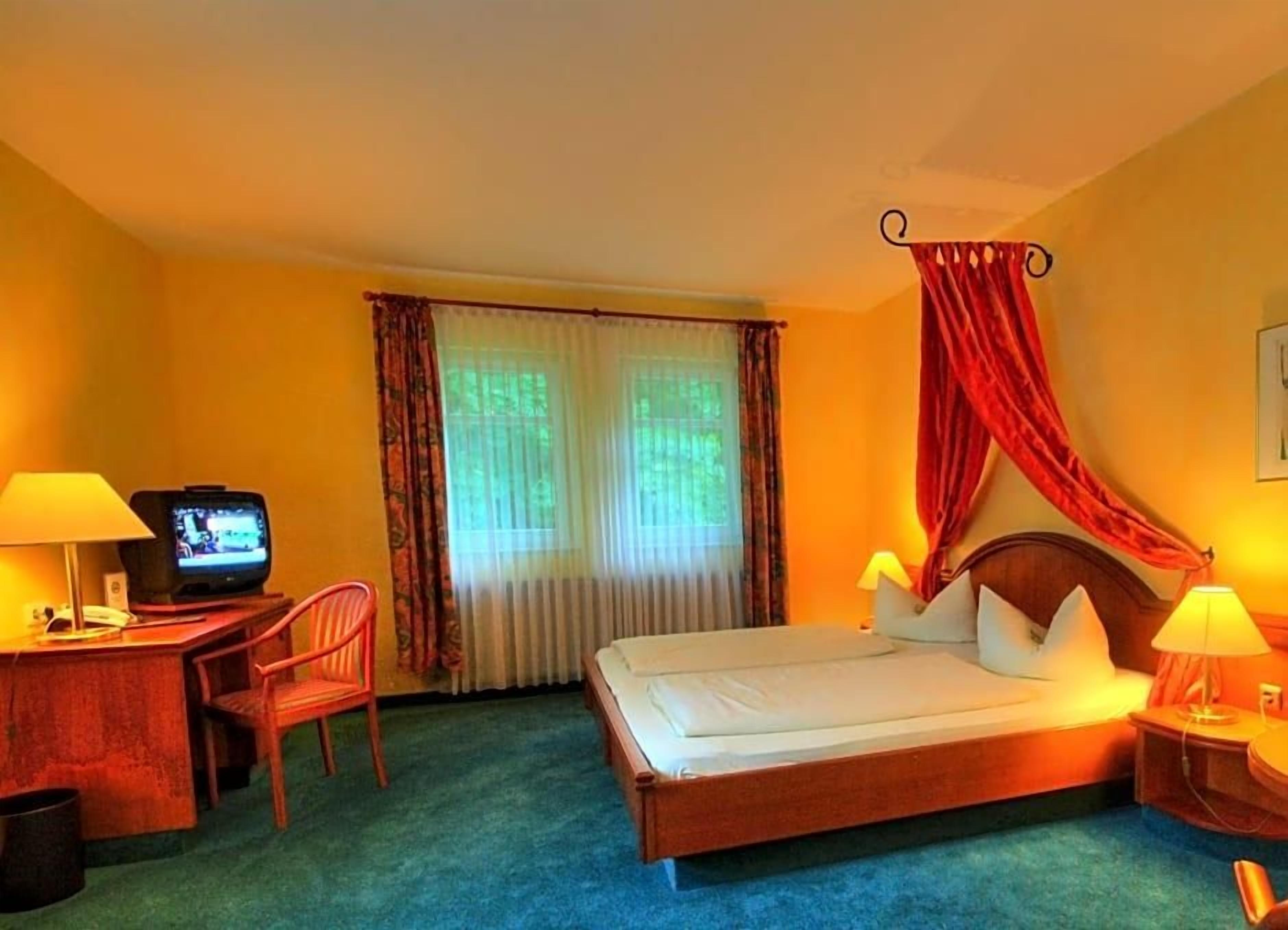 Ein Hotelzimmer mit einem Bett, einem Schreibtisch mit Fernseher, einem Sessel und einem Fenster mit Vorhängen.