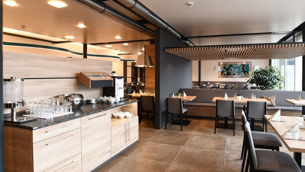 A modern restaurant with a central counter, seating area, and a large window. - WESTSIDE Hotel Garni (Munich)