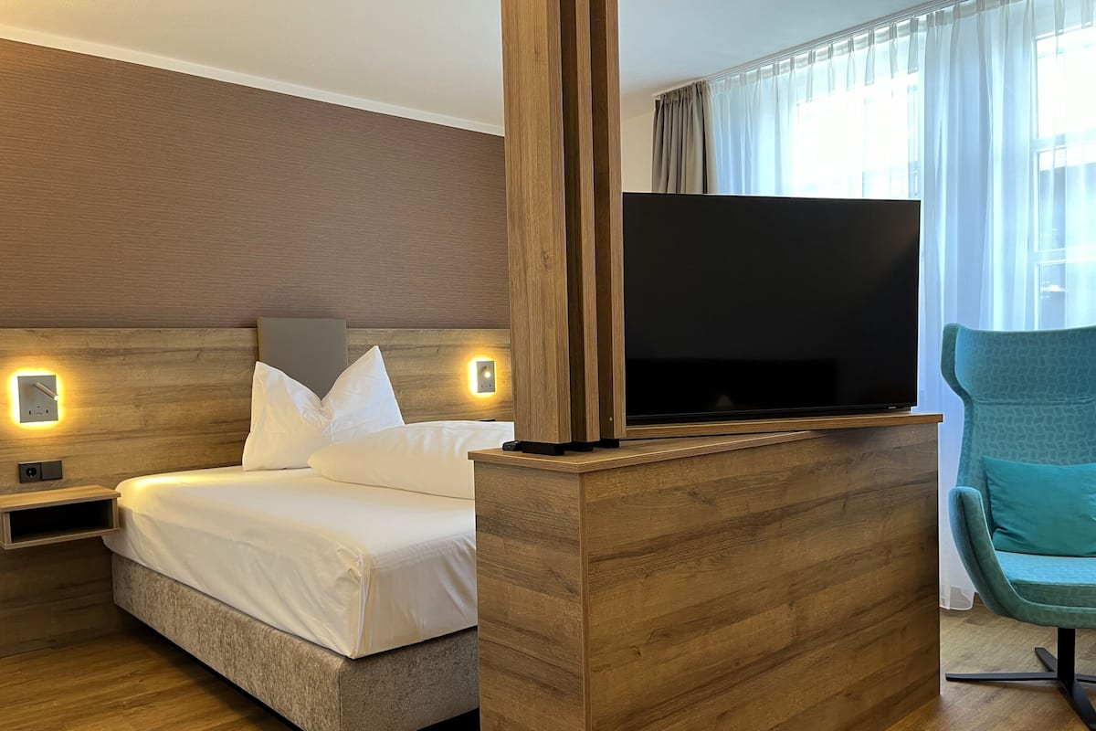 Une chambre dâhĂŽtel avec un lit, un tĂ©lĂ©viseur posĂ© sur un meuble en bois, une table de chevet et un fauteuil de couleur turquoise.