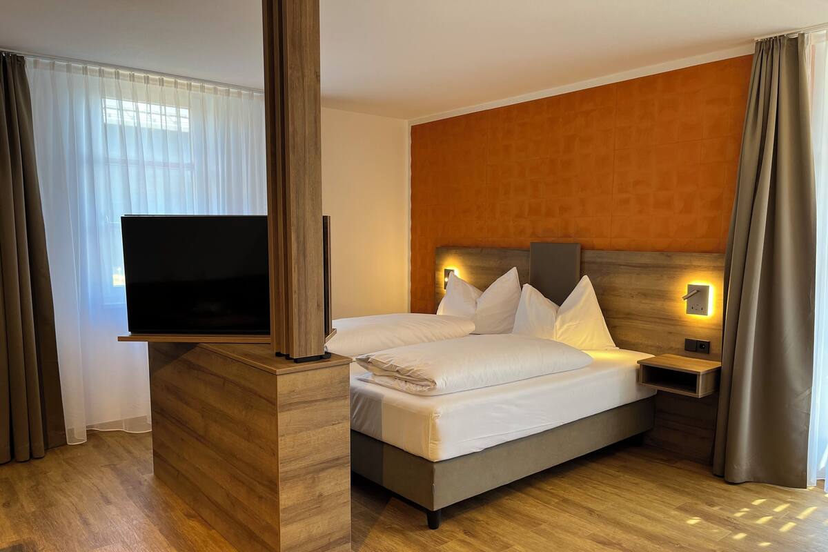 Une chambre dâhĂŽtel moderne avec une tĂȘte de lit en bois, un tĂ©lĂ©viseur Ă Ă©cran plat, un lit avec du linge de lit blanc et un plancher en bois.
