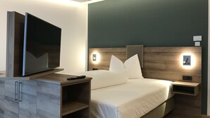 A modern hotel room with a large flat-screen TV on a wooden cabinet, a neatly made bed with white linens, and built-in wall sconces. - WESTSIDE Hotel Garni (Munich)