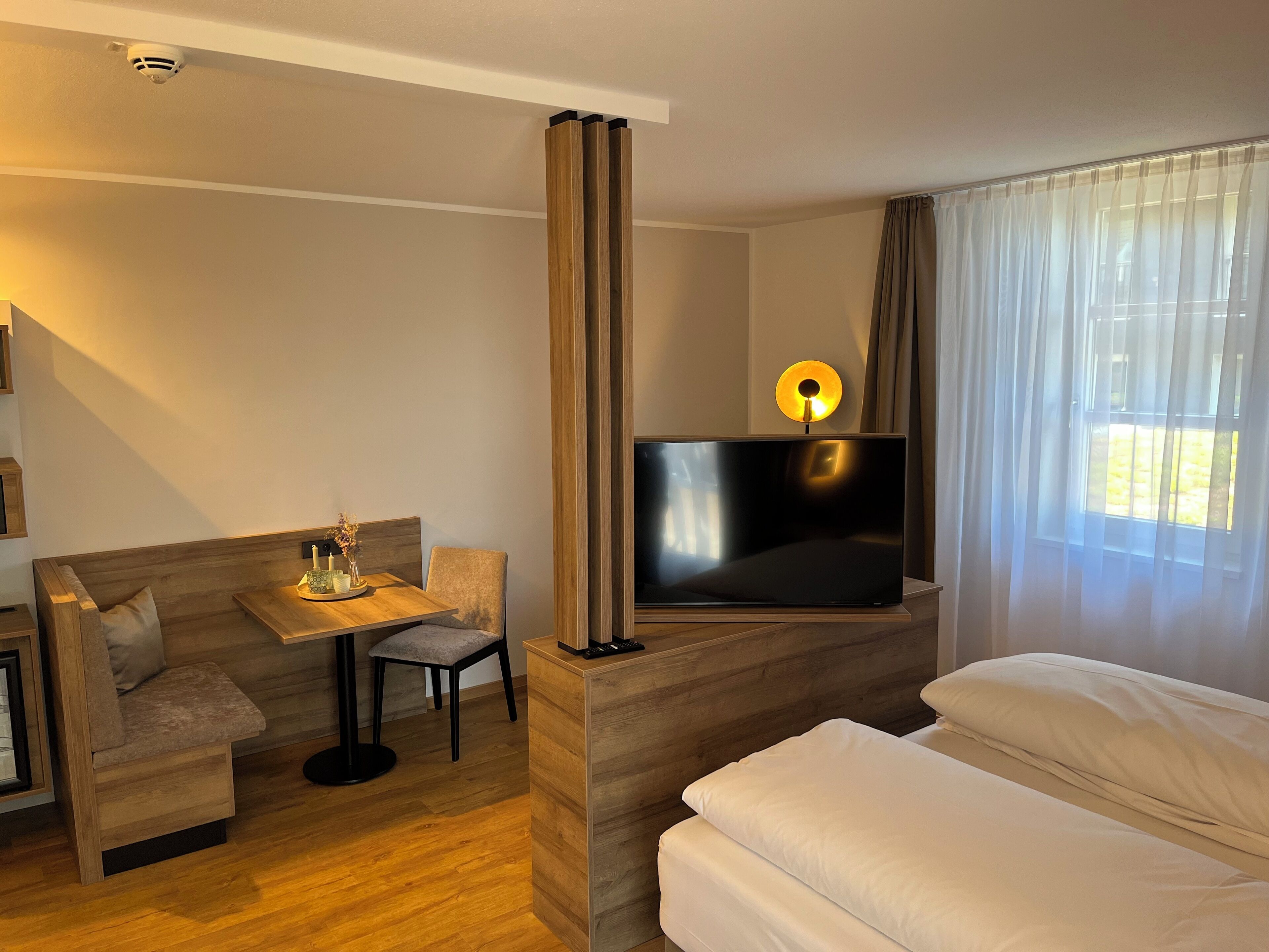 Habitación de hotel con un sofá de madera, una mesita con velas, un televisor sobre un mueble de madera, una cama con sábanas blancas y una ventana con cortinas translúcidas.