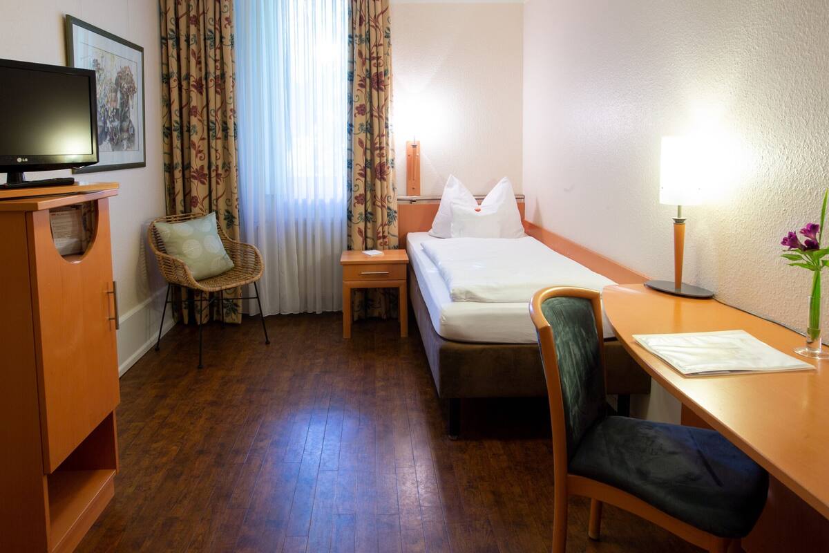 Une chambre d’hôtel avec un lit, un bureau avec une lampe, une chaise, une télévision et une fenêtre avec des rideaux.