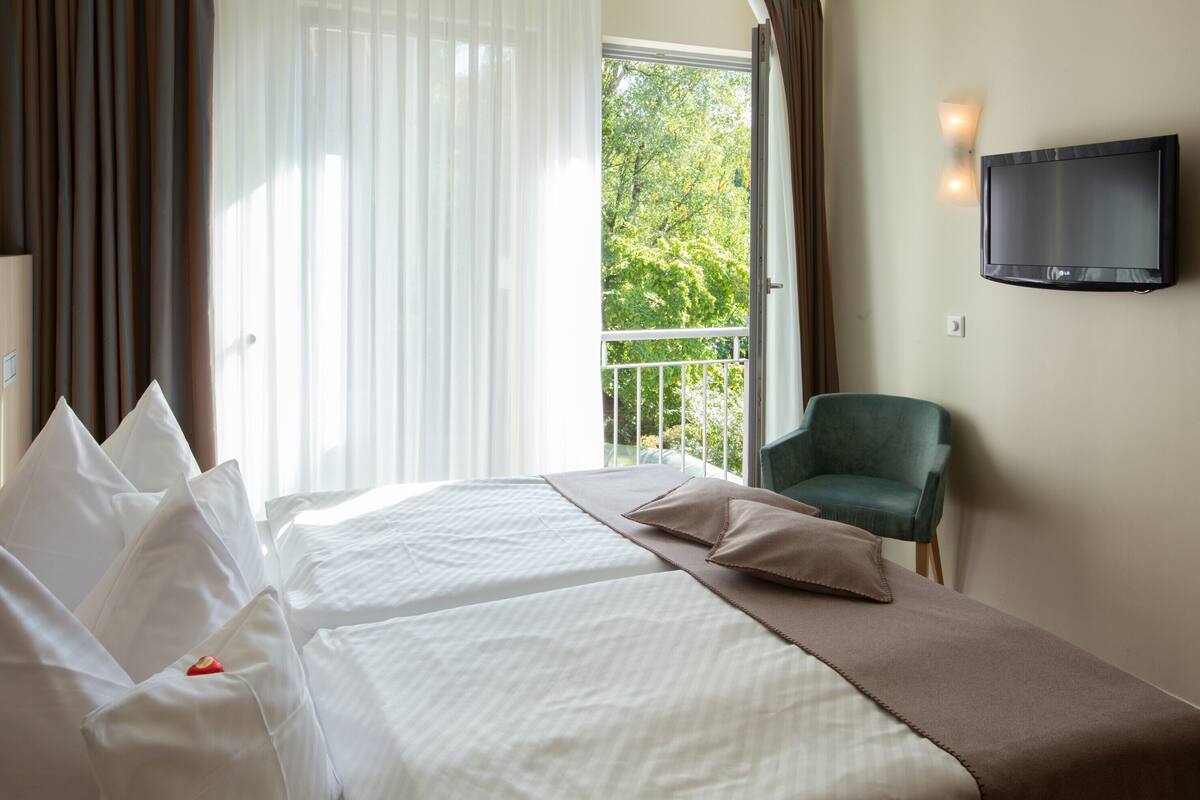 Une chambre d’hôtel avec un lit, un fauteuil vert, une télévision et un balcon donnant sur des arbres.