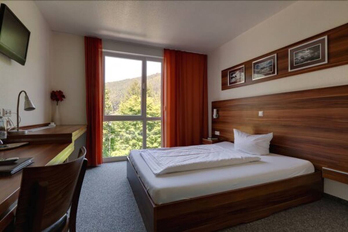 A hotel room with a wooden headboard, a bed with white linens, a desk with a lamp, and a window with curtains.