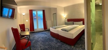 Quarto de hotel com cama de casal, escrivaninha com cadeira, televisão na parede e porta de correr para uma varanda.