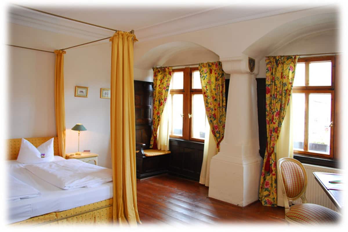 A room with a four-poster bed, a desk with a chair, and two windows with floral curtains.