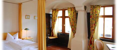 A room with a four-poster bed, a desk with a chair, and two windows with floral curtains.