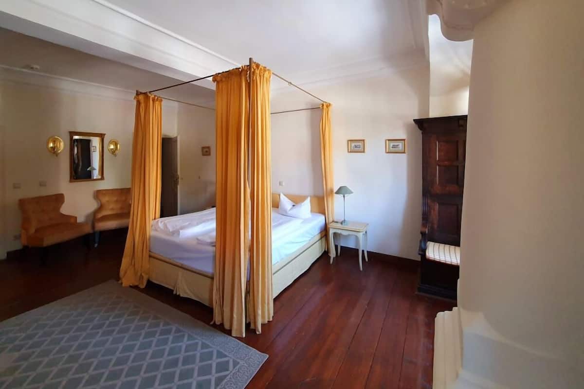 A bedroom with a four-poster bed, a small bedside table with a lamp, a chair, and a piano.