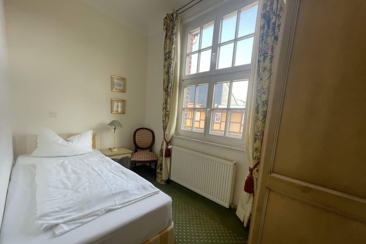 A hotel room with a bed, a chair, a table with a lamp, a window with curtains, and a radiator.