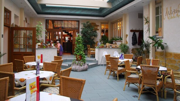A restaurant with wicker furniture, a central plant, and a view of the kitchen in the background. - Hotel Adler (Rudolstadt)