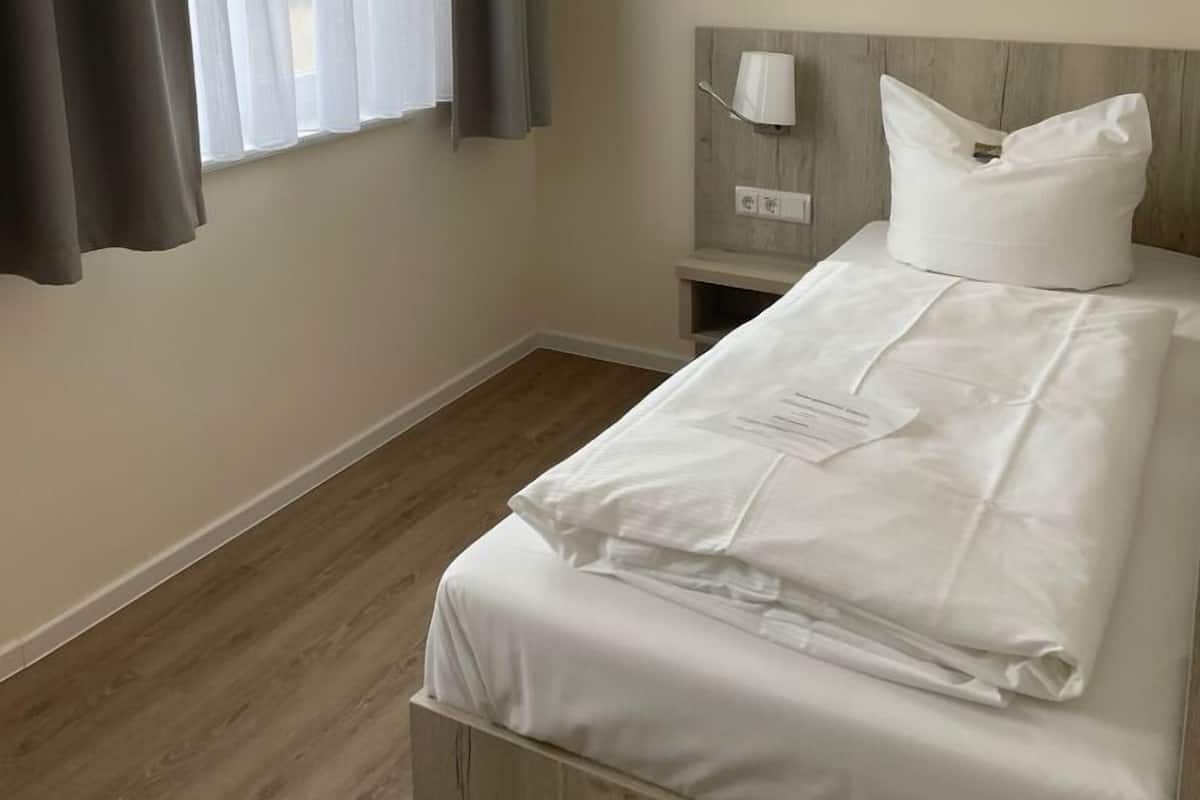 A neatly made bed with white linens, a bedside table with a lamp, and a window with sheer curtains.