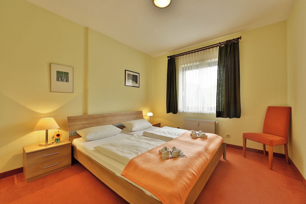 A hotel room with a bed, a chair, a nightstand with a lamp, and two framed pictures on the wall.