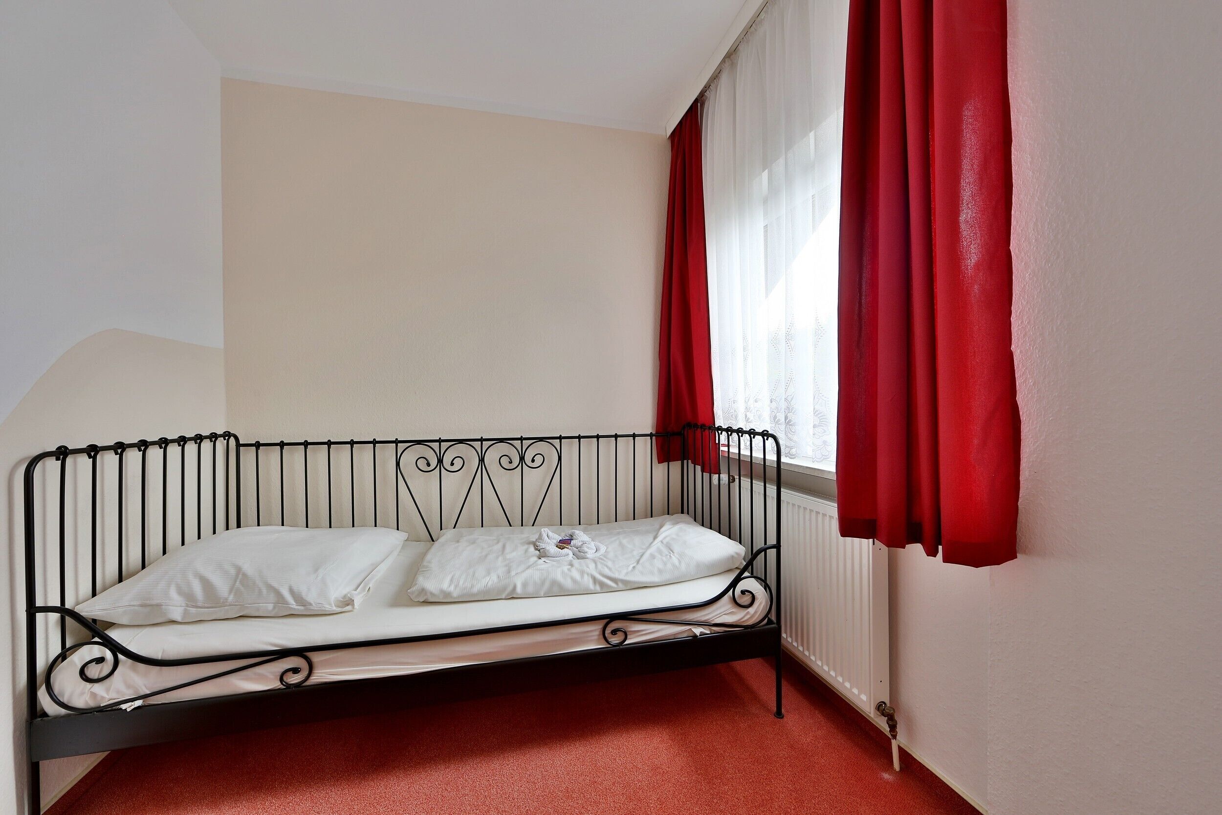 A single bed with a metal frame, white bedding, and red curtains.