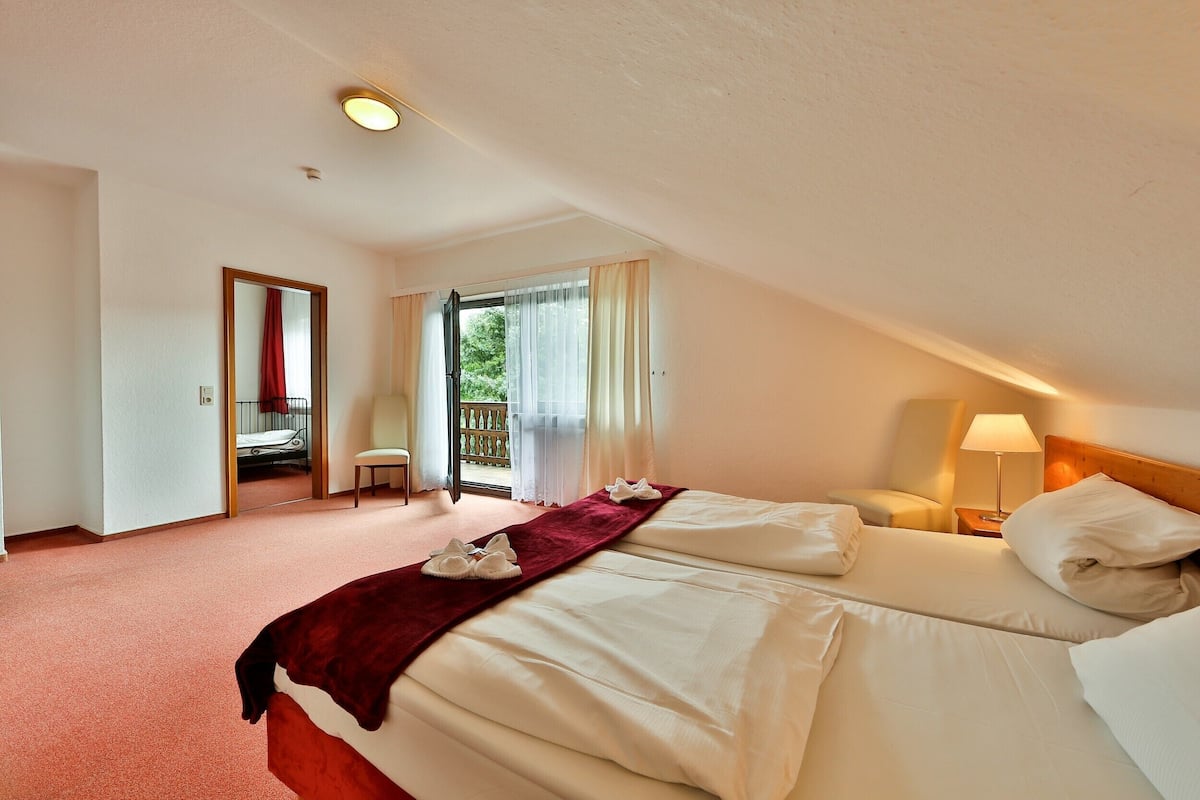 A hotel room with two beds, a red carpet, a wooden headboard, a nightstand with a lamp, and a view of the outdoors through a door.