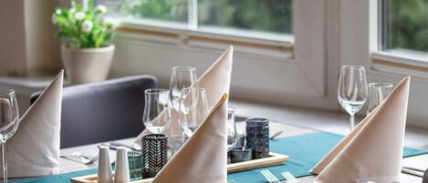 Une table à manger dressée avec des serviettes blanches, des couverts en argent et de la verrerie, et une vue sur la verdure à travers une fenêtre.