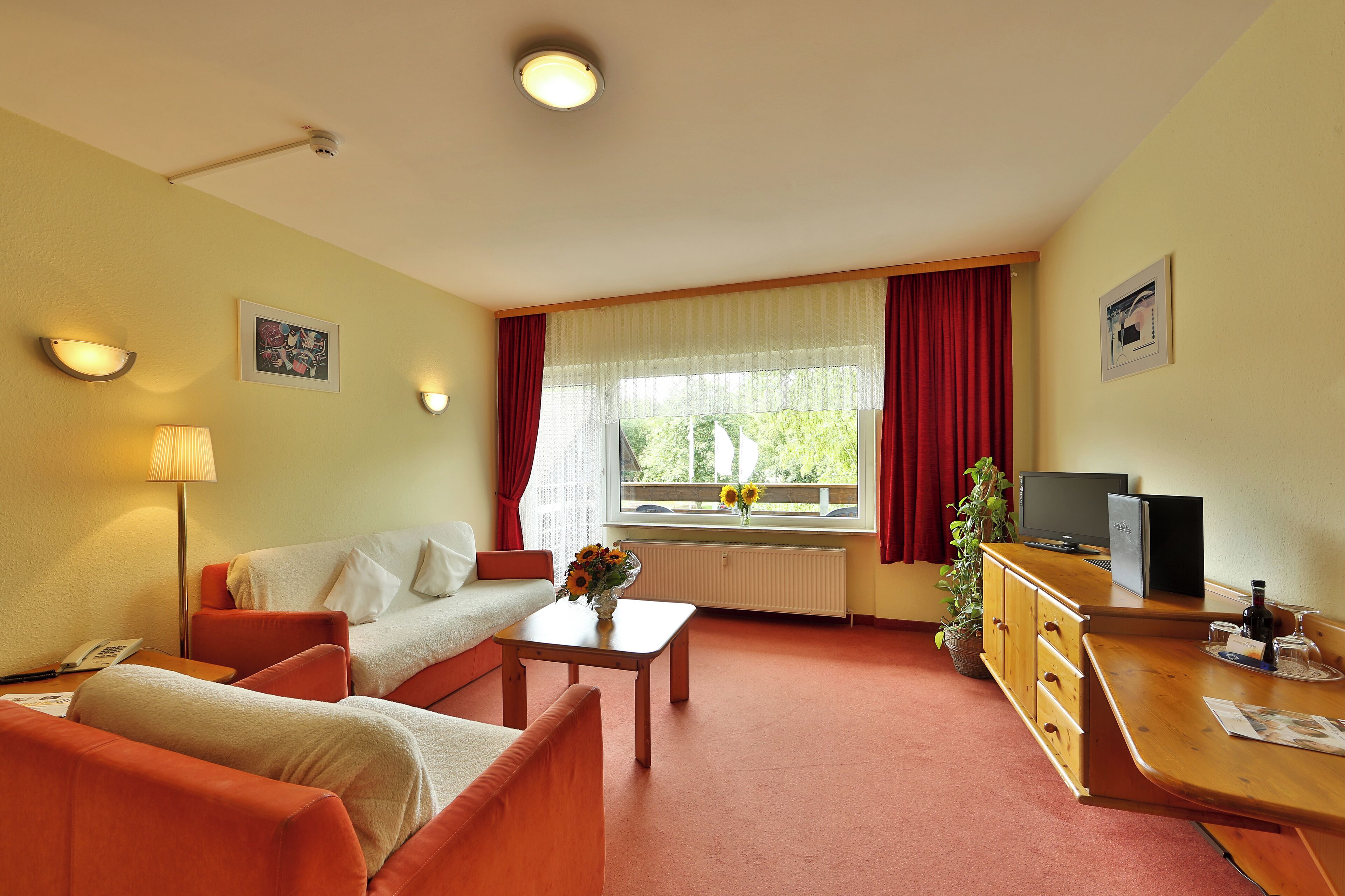 Een hotelkamer met een bank, een tafeltje met een bloemenvaas, een bureau met een computer, een televisie en een raam met gordijnen.