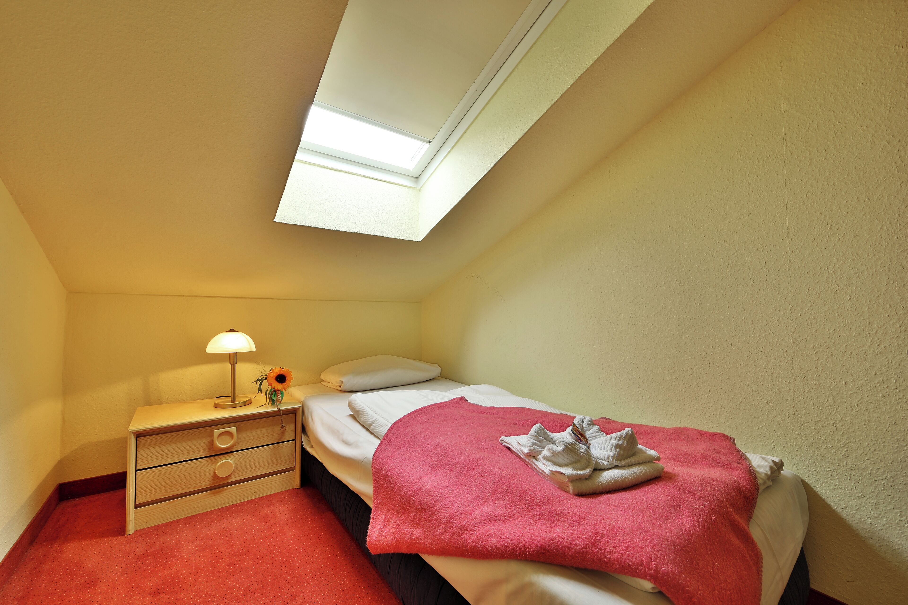 Une petite chambre à coucher, bien rangée, avec un lit simple, une table de chevet, une lampe et un puits de lumière.