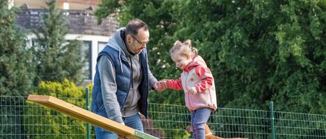 A man and a child on a seesaw in an outdoor area with a green fence and trees in the background.