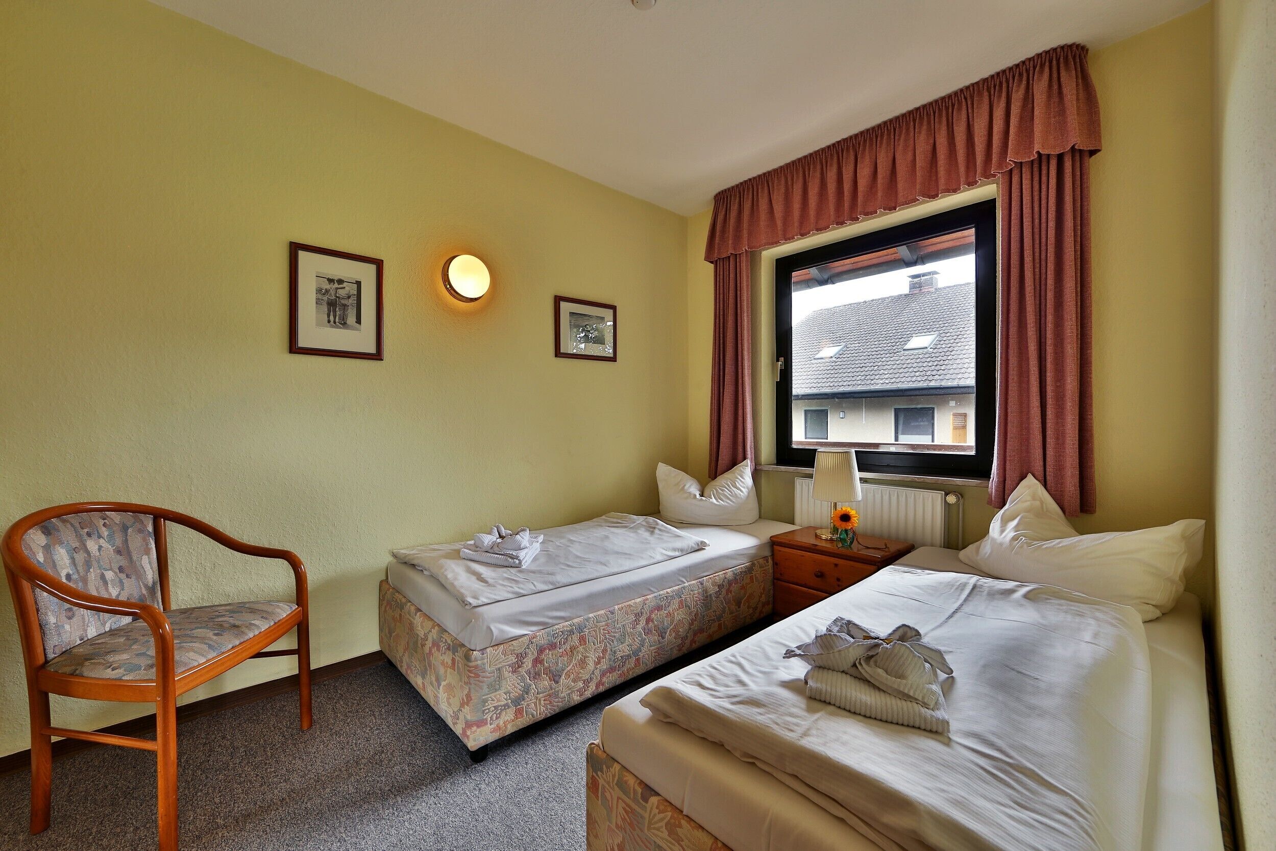 Habitació d'hotel amb dos llits individuals, una cadira de fusta, una taula petita amb una làmpada i una finestra amb cortines.