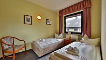 Habitació d'hotel amb dos llits individuals, una cadira de fusta, una taula petita amb una làmpada i una finestra amb cortines.
